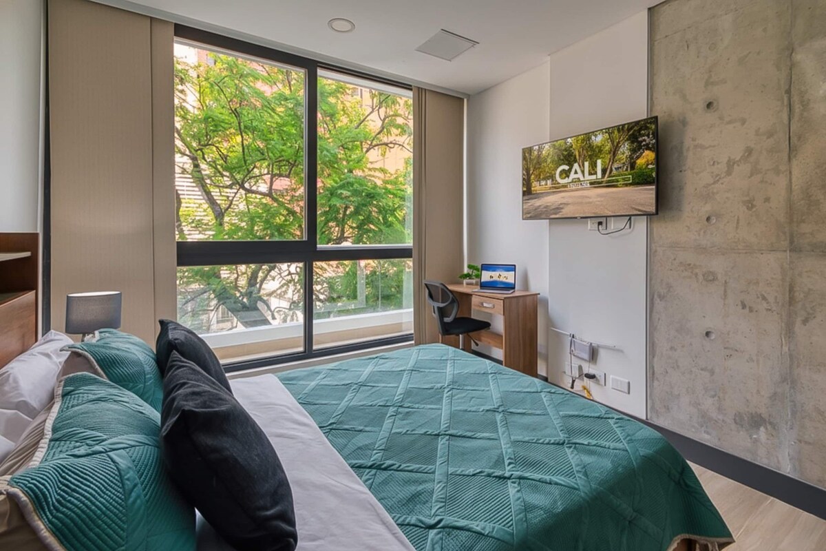 A hotel room with a large window, a bed with green bedding, a desk with a computer, and a TV mounted on the wall.