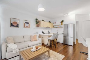 A compact living space with a sofa, coffee table, kitchen area, and a refrigerator. - Courtyard 1-Bed Apartment in New Farm (New Farm)
