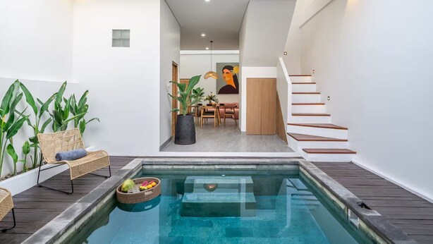 A modern indoor pool area with a wooden deck, lounge chairs, and a dining area with a table and chairs. - Kutesa Villa Bali (Kerobokan)