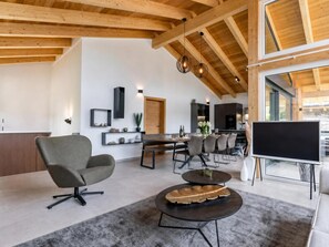 Ein modernes Wohnzimmer mit hohen Decken, Holzbalken, einem Flachbildfernseher, einer Essecke mit Tisch und Stühlen sowie einer Küche im Hintergrund.