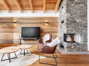 Ein modernes Wohnzimmer mit Kamin aus Stein, Holzbalkendecke, Flachbildfernseher, Ledersessel und runden Couchtischen.