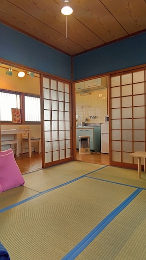 Ein traditioneller japanischer Raum mit Tatami-Böden, Schiebetüren, einer Küche im Hintergrund und einem kleinen Tisch mit Stuhl.