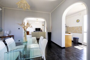 Ein modernes Wohnzimmer mit einem Glastisch, weißen Stühlen, einem Fernseher und einer durch einen Bogen sichtbaren Küche.