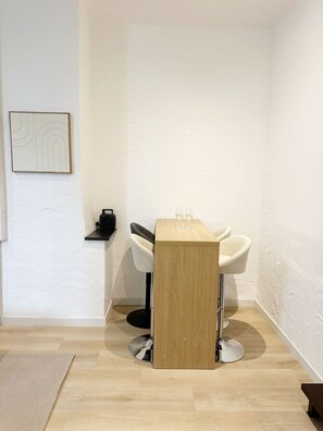 Une pièce d’angle avec une table en bois, deux chaises blanches, une petite étagère sur laquelle se trouve une machine à café, et un tableau encadré accroché au mur.