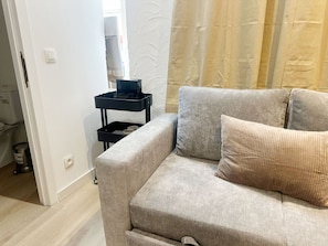 A living room with a beige sofa, a black side table with a television, and a window with curtains. - Cozy 1BR & Living Room Near City Center (Ghent)