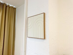 Une pièce avec un rideau beige, un mur blanc et une œuvre d’art encadrée.