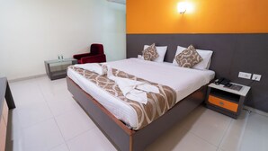 A modern hotel room with a large bed, a bedside table with a phone, a red chair, and a nightstand with a lamp. - PLA Residency Tanjore (Thanjavur)