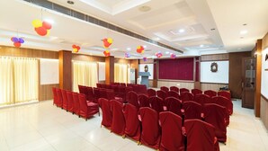 A conference room with rows of red chairs, a podium, a screen, and decorative balloons. - PLA Residency Tanjore (Thanjavur)