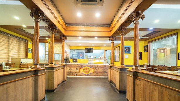 A restaurant interior with wooden counters, a central counter with a kebab station, and a ceiling with recessed lighting. - PLA Residency Tanjore (Thanjavur)