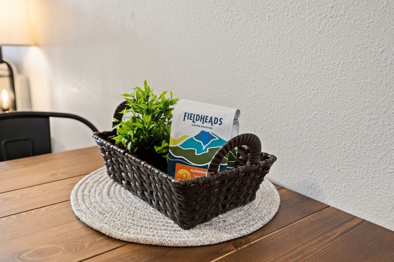 Een rieten mand met een koffiezakje en een plantje op een ronde geweven onderzetter op een houten tafel.