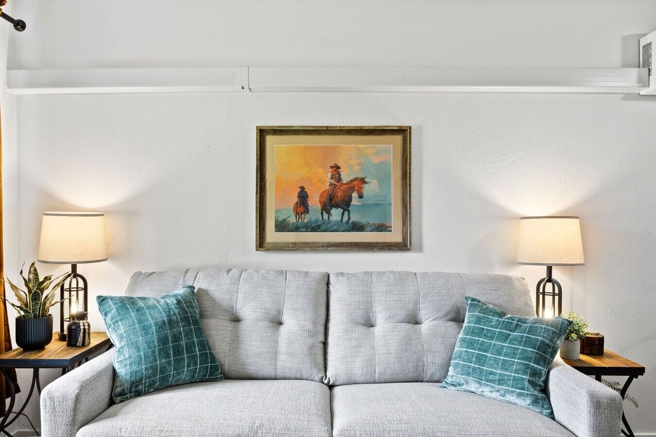 A living room with a grey sofa, teal pillows, two side tables with lamps, and a framed painting on the wall.
