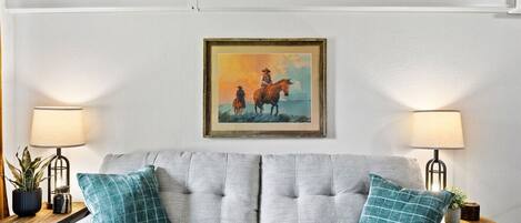 A living room with a grey sofa, teal pillows, two side tables with lamps, and a framed painting on the wall.