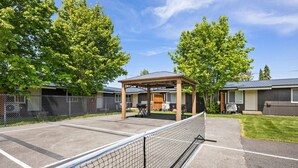 A fenced tennis court with a covered seating area, a building with a porch, and a grassy area with trees. - Family Stay Near Glacier + Whitefish w/ Pickleball (Columbia Falls)