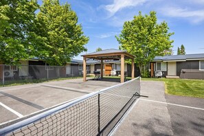 Una cancha de tenis cercada con una zona de estar cubierta, un edificio con porche y un área verde con árboles.