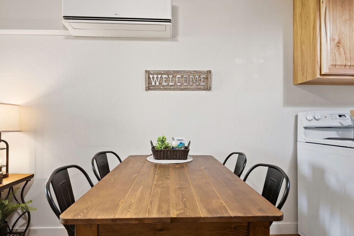 A wooden dining table with four chairs, a welcome sign, a basket with plants, and a white oven.