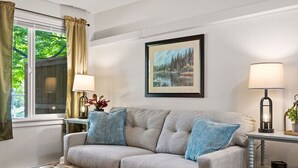 A living room with a sofa, two lamps, a framed painting, and a window with curtains. - Glacier NP Retreat w/ Modern Comforts & Pickleball (Columbia Falls)