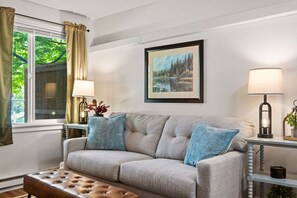 Un salon avec un canapé, deux lampes, un tableau encadré et une fenêtre avec des rideaux.