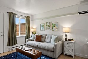 Ein Wohnzimmer mit grauem Sofa, einem braunen Hocker, einem Beistelltisch mit Lampe und einem Fenster mit Vorhängen.