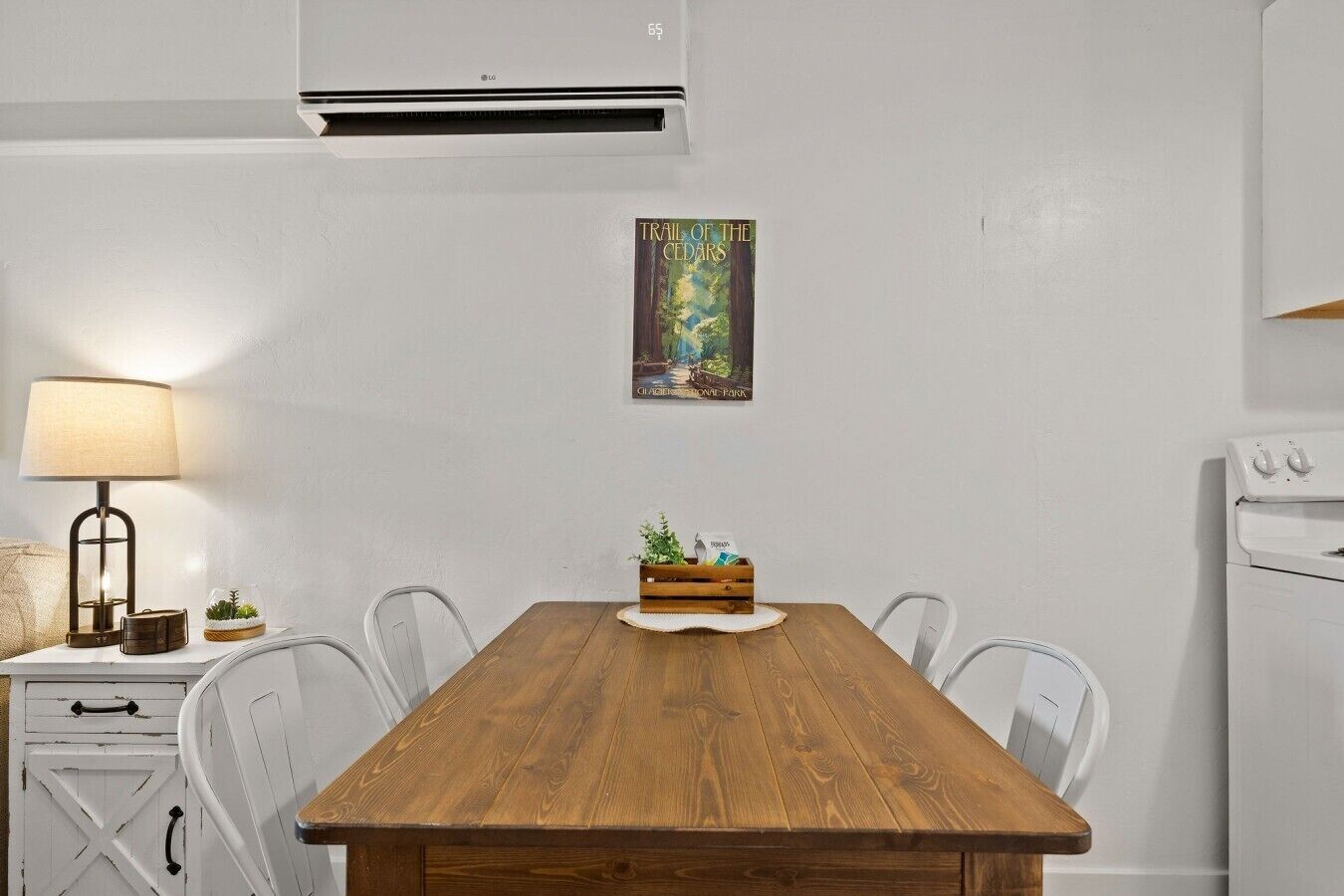 A dining area with a wooden table, chairs, a lamp, a small plant, and a framed picture on the wall.