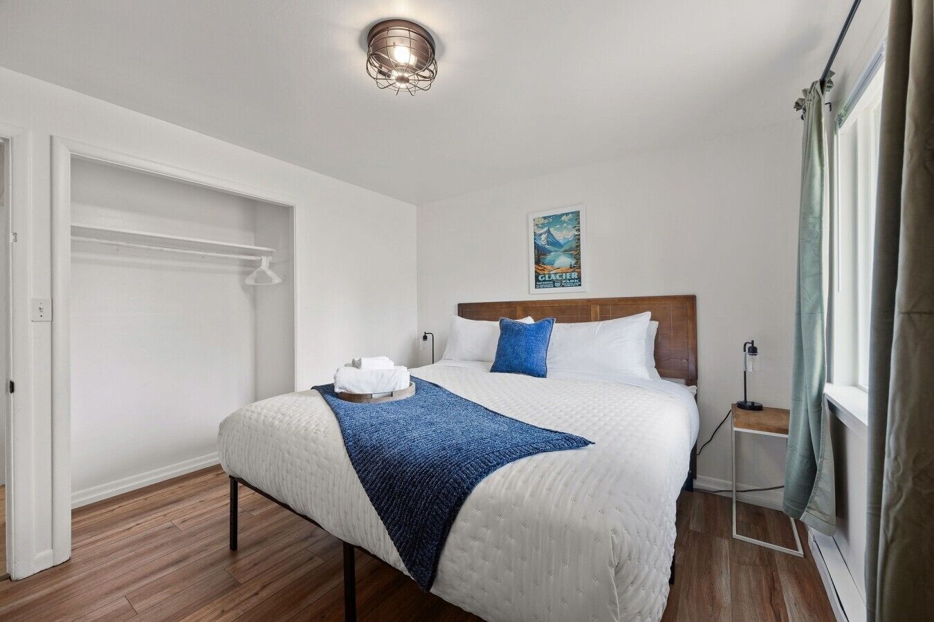 Une chambre d’hôtel avec un lit, une table de chevet, une fenêtre avec des rideaux, un luminaire fixé au mur et une affiche accrochée au mur.