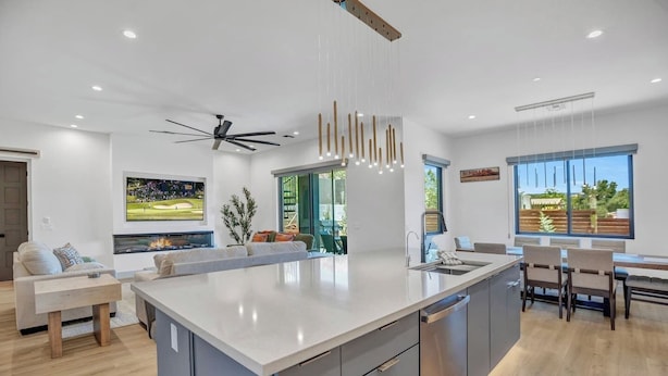 Una cocina moderna con una isla central, un área de comedor con mesa y sillas, un ventilador de techo, iluminación empotrada y un ventanal con vistas.