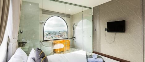 Una habitación de hotel moderna con una cama grande, cabina de ducha acristalada, televisor y vistas a la ciudad.