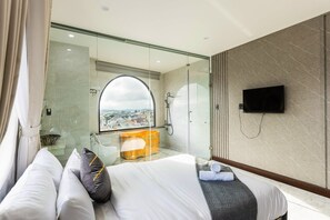 Quarto de hotel moderno com uma cama grande, chuveiro em cabine de vidro, televisão e vista para a cidade.