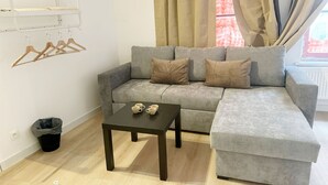A grey sofa with two pillows, a small table with two cups, wooden hangers on a wall-mounted rack, a black trash bin, and a window with curtains. - Convenient Studio in the Heart of City (Ghent)