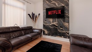 A modern living room with a leather sofa, a flat-screen TV mounted on a wall with a black and gold abstract design, and a window with blinds. - 4 bedroom house,  sleeps 8, fast WIFI, shops, long stays, pet friendly (Nelson)