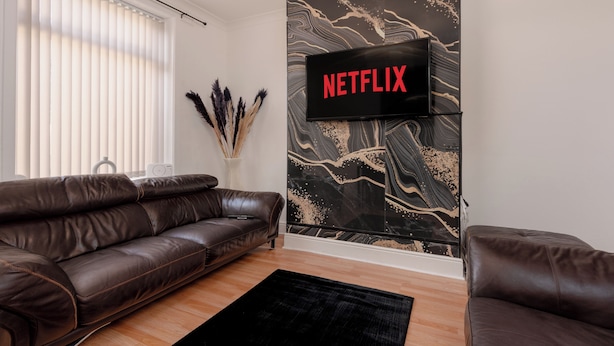 Ein modernes Wohnzimmer mit einer Ledersofa, einem flachbildfernseher an der Wand, der mit einem schwarz-goldenen abstrakten Muster versehen ist, und einem Fenster mit Jalousien.