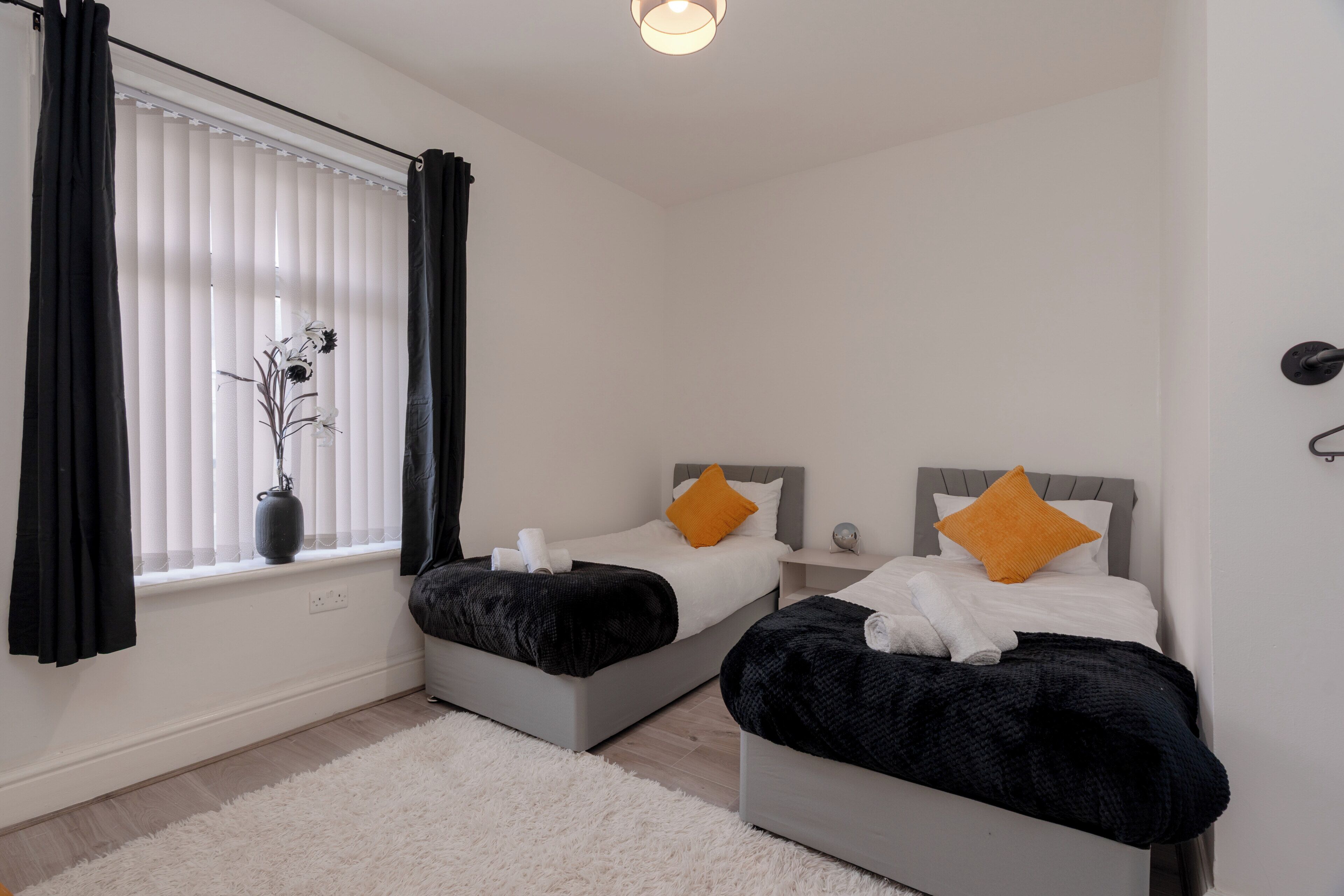 A bedroom with two beds, a window with blinds, a bedside table, and a white rug.
