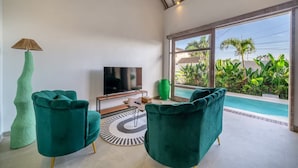 A modern living room with a TV, two green chairs, a lamp, and a view of a swimming pool. - La Palma Villa Bali (Kerobokan)