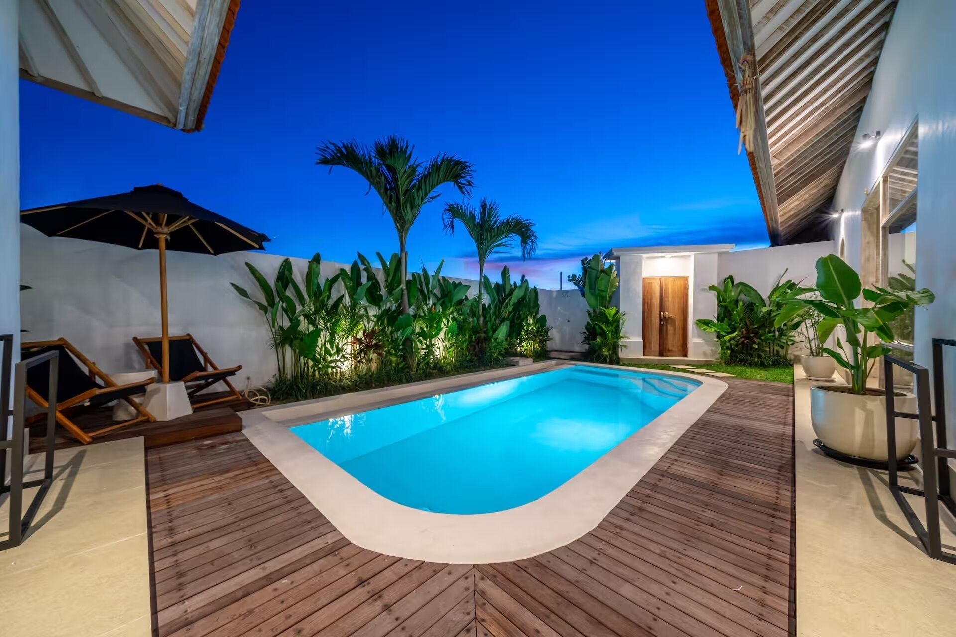 A swimming pool with wooden decking, surrounded by tropical plants and a palm tree, under a clear sky.