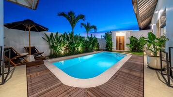 A swimming pool with wooden decking, surrounded by tropical plants and a palm tree, under a clear sky.