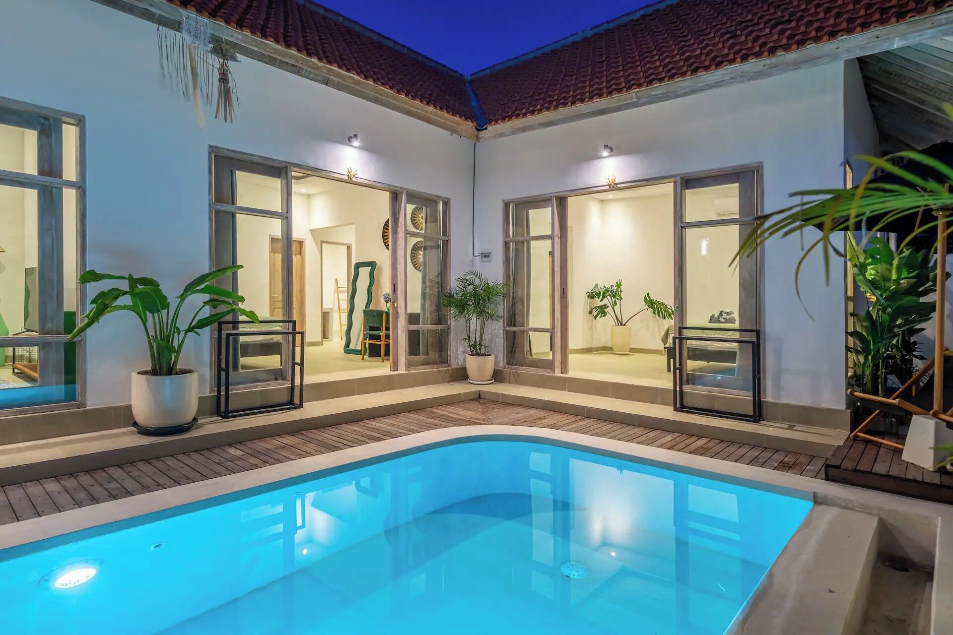 A swimming pool with glass doors leading to a room, surrounded by potted plants and a wooden deck.