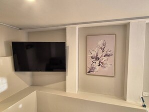 Ein Zimmer mit einem an der Wand befestigten Flachbildfernseher, einer gerahmten Blumenillustration und einem weißen Regal.