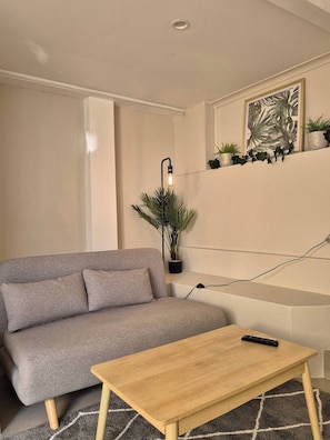 Ein Wohnzimmer mit grauem Sofa, einem Holztisch, einem Blumentopf, einer Wandleuchte und einem gerahmten Bild.