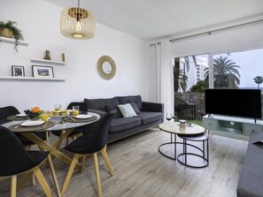Ein modernes Wohnzimmer mit grauem Sofa, Esstisch, Stühlen, Fernseher und einem großen Fenster mit Blick auf Palmen.