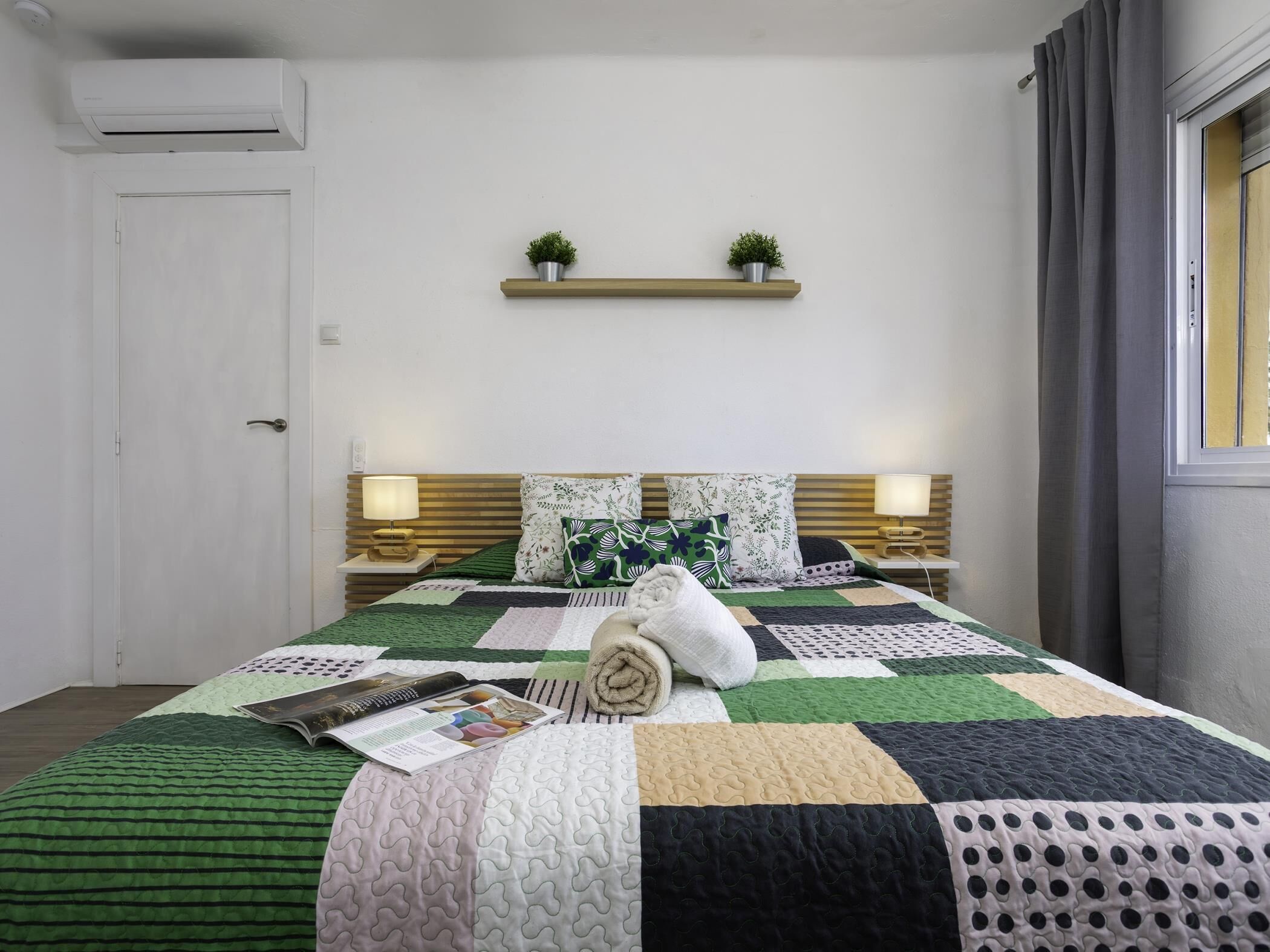 Een netjes ingerichte slaapkamer met een bed, nachtkastjes, een plank met planten en een airconditioning.