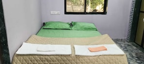 Un lit simple avec une couvre-lit verte et deux oreillers, une fenêtre donnant sur un paysage verdoyant et une prise électrique sur le mur.