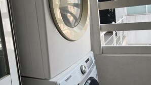 Une laveuse et une sécheuse Samsung à chargement frontal, superposées dans une buanderie avec une fenêtre et une rambarde en métal.