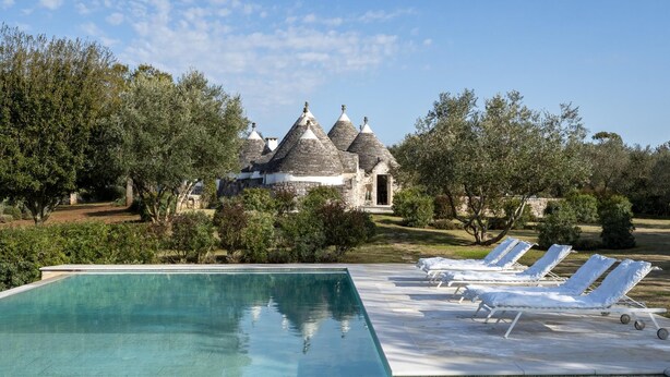 Ein Pool mit Liegestühlen, ein Steinhaus mit Kegeldächern und umliegendes Grün.