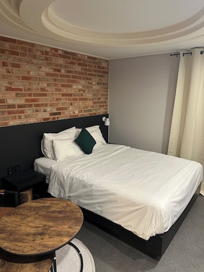 Una habitación de hotel con pared de ladrillo, una cama con ropa blanca, una mesa redonda de madera y una mesita de noche.