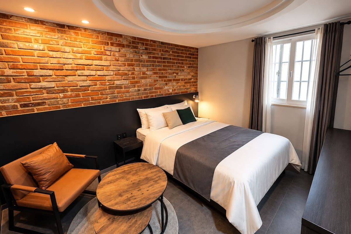 Habitación de hotel con pared de ladrillo, cama con ropa de cama blanca, mesa de madera redonda, sofá y mesita de noche.