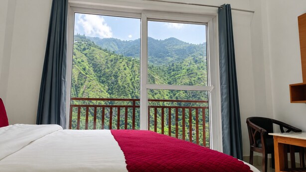 A hotel room with a bed, a wooden desk, and a balcony with a view of a forested mountain.