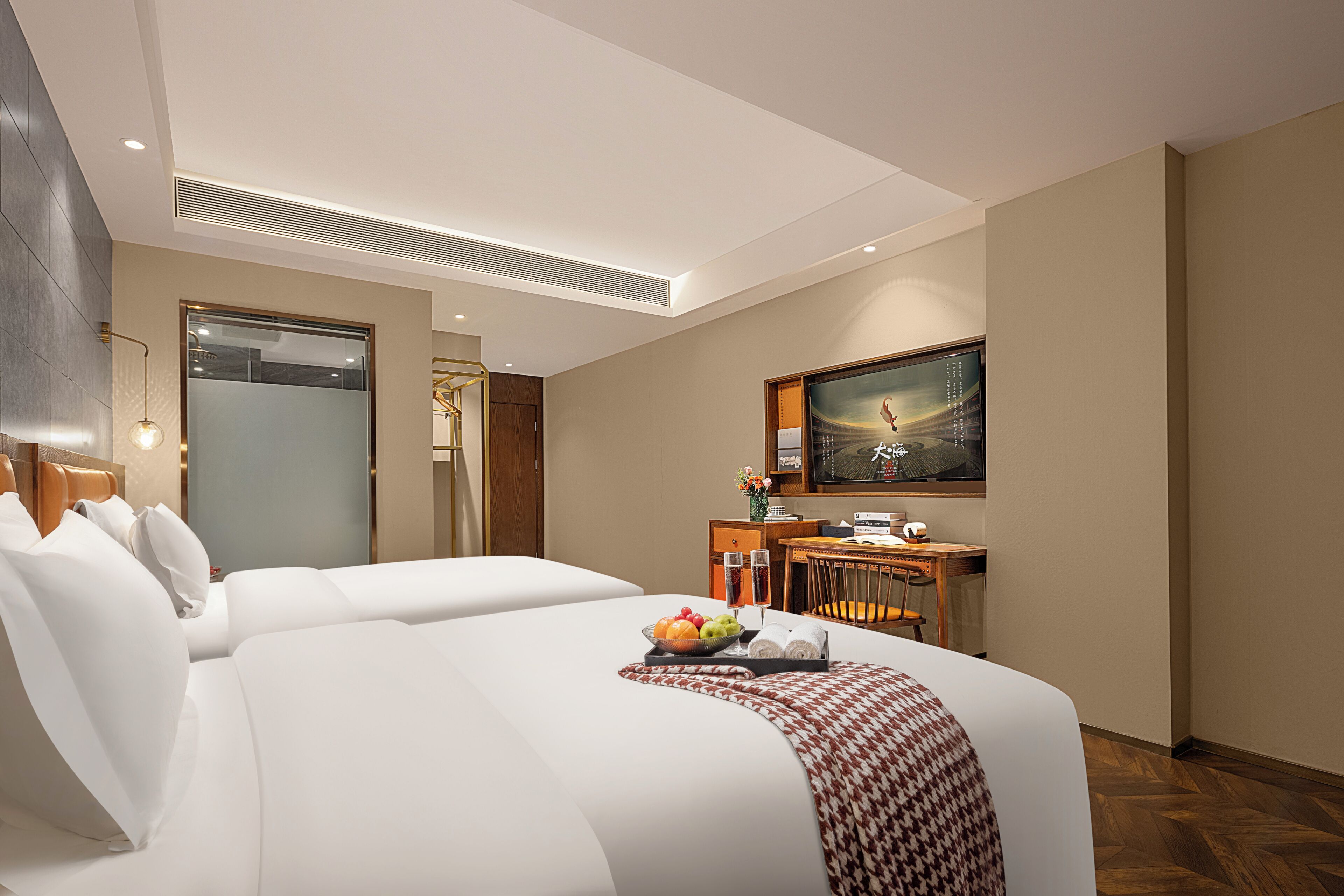 Une chambre d’hôtel avec deux lits, un bureau, une chaise, une télévision et un panier de fruits posé sur l’un des lits.