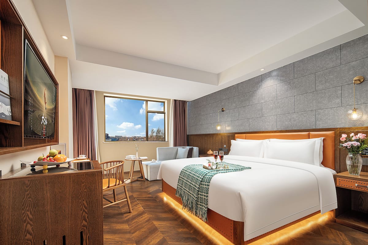Habitación de hotel con una cama grande, un escritorio con frutero, una mesita, una silla y vistas al cielo a través de una ventana.