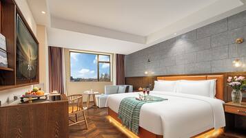 A hotel room with a large bed, a desk with a fruit bowl, a small table, a chair, and a view of the sky through a window.