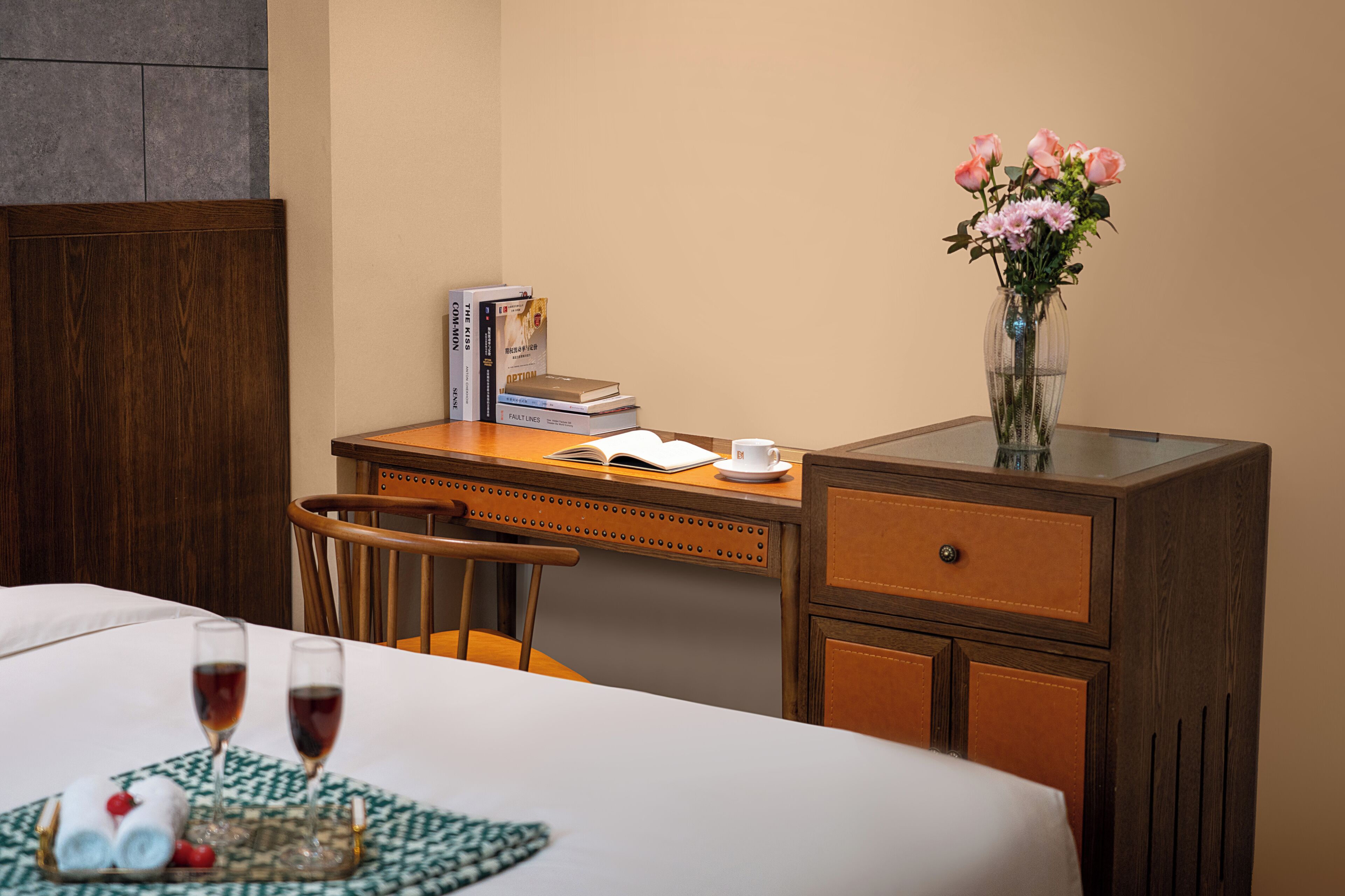 A hotel room with a bed, a wooden desk with books, a vase with flowers, and two glasses of red wine.