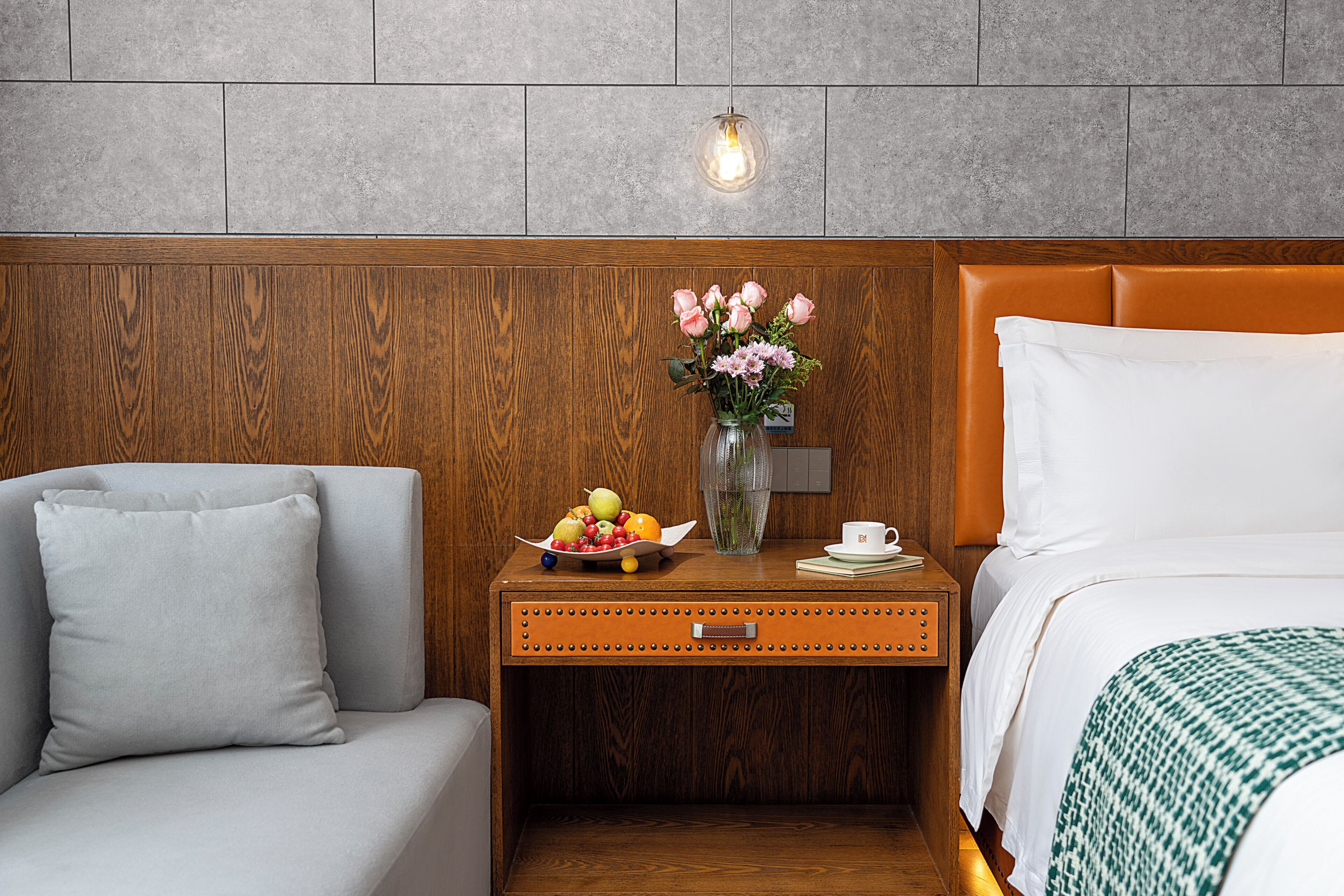 A hotel room with a wooden headboard, a nightstand with a vase of flowers, a fruit bowl, and a bed with a green patterned blanket.